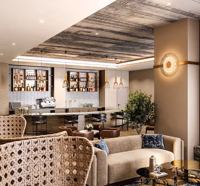 A modern and stylish lounge area featuring a beige sofa with blue patterned pillows, woven chairs, a small round coffee table, and a bar counter with high stools. The bar shelves are stocked with bottles and glassware, illuminated by pendant lights hanging from a textured ceiling. There is a green potted plant near the bar area and a decorative wall light on a beige column.