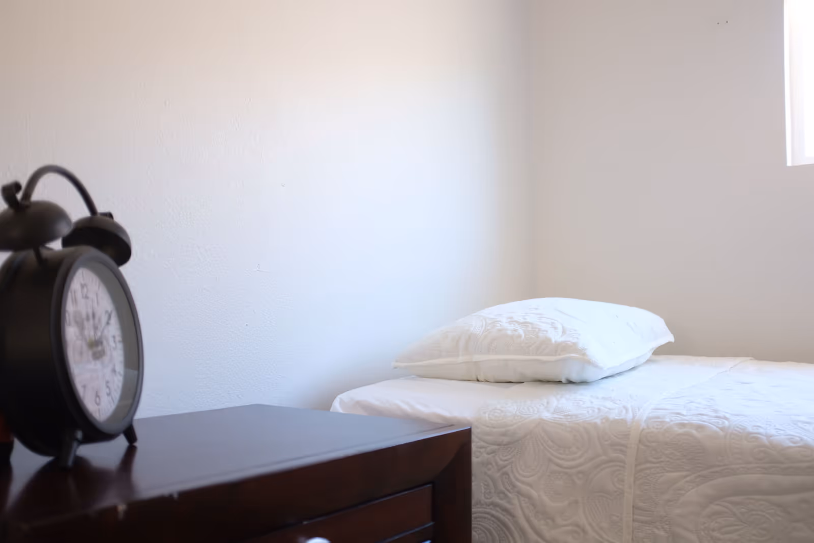 Simple bedroom with a single bed dressed in white bedding and a pillow, a wooden nightstand, and a black alarm clock.