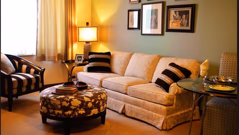 A cozy living room with a beige sofa adorned with striped black and beige pillows, a round floral-patterned ottoman with books on top, a striped armchair, a side table with a lamp and framed photos, and a glass-top dining table with a place setting. The walls are decorated with framed artwork and the room is softly lit by natural light from a window with beige curtains.