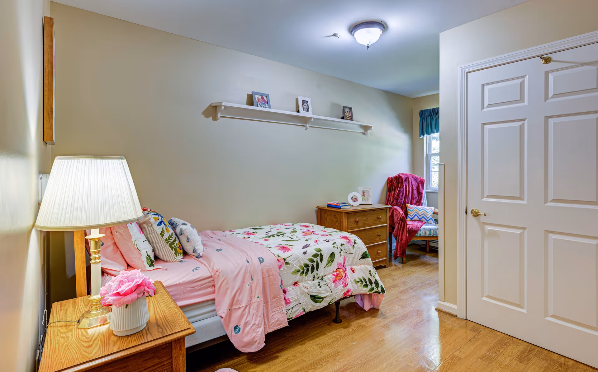A cozy bedroom with a single bed covered in pink floral bedding and multiple pillows. A wooden nightstand with a lamp and a pink flower vase is beside the bed. On the opposite side, there is a wooden dresser with picture frames and books on top. A comfortable chair with a red throw blanket and a colorful cushion is placed near a window with blue curtains. The room has wooden flooring and beige walls with a white door on the right side.