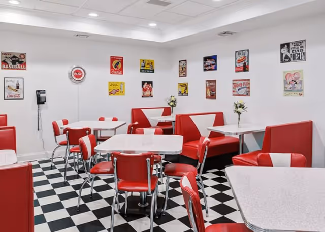 A retro-style dining room with red and white booths and chairs, white tables, and a black and white checkered floor. The walls are decorated with vintage-style posters and signs, including Coca-Cola and Elvis Presley themes. There are small flower vases on some tables.