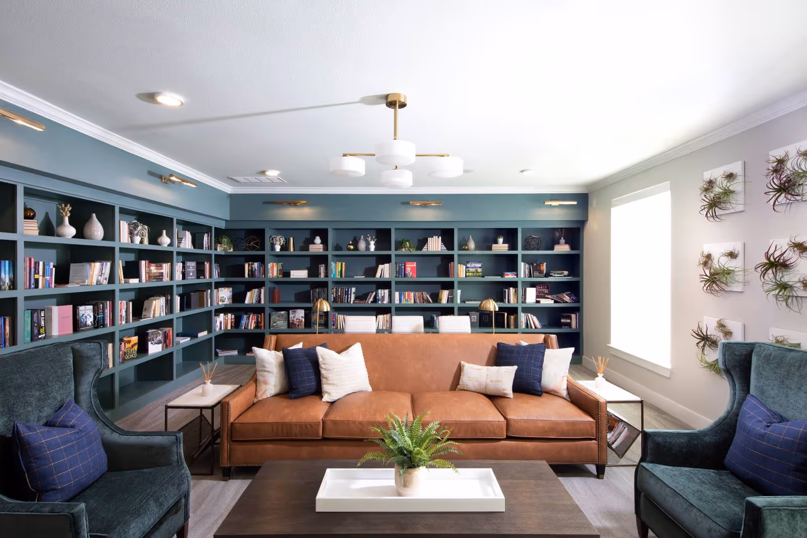 Bright communal living room with a tan leather sofa, green armchairs, a coffee table, and built-in teal bookshelves filled with books and decor.
