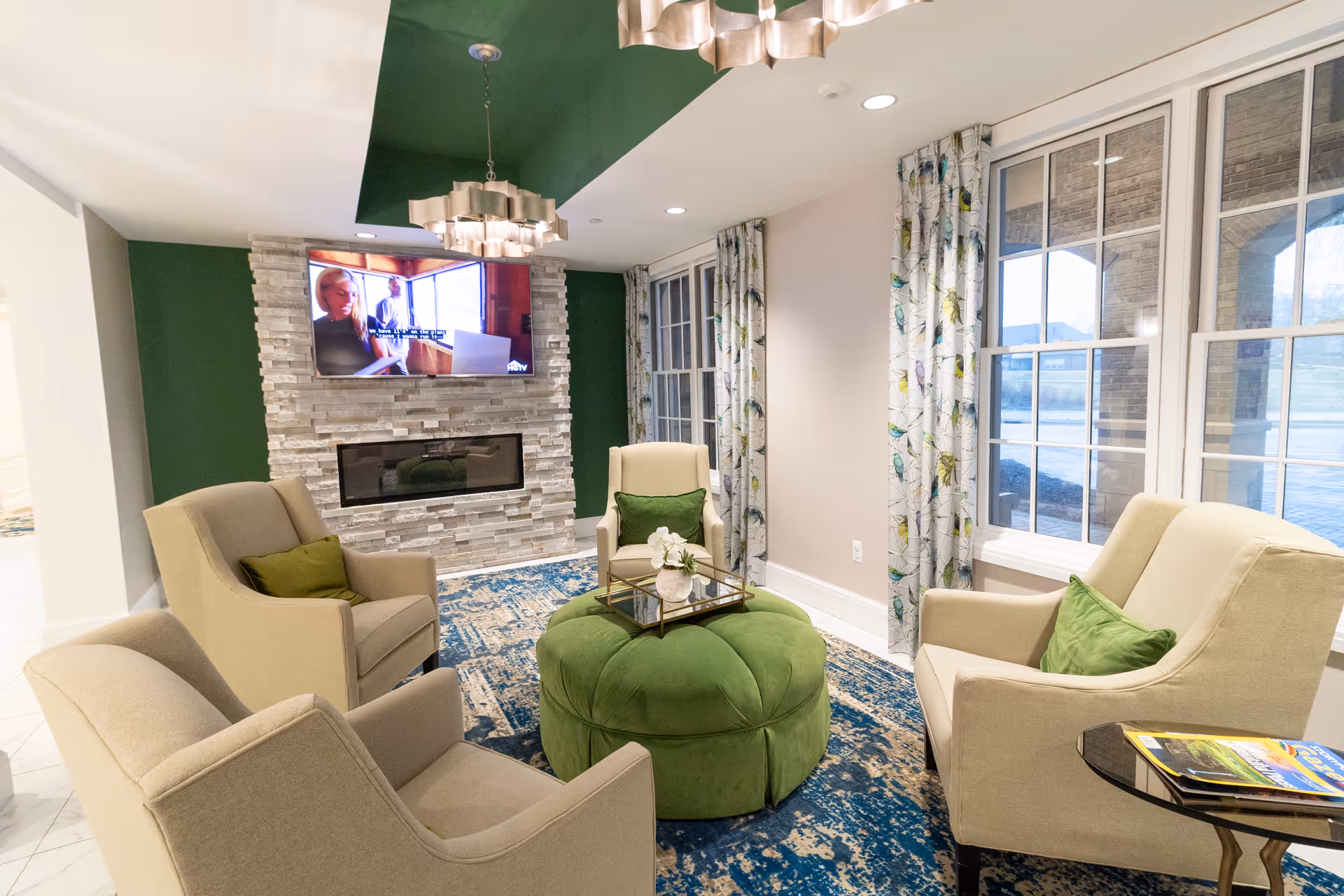 A cozy living room area with four beige armchairs arranged around a large green ottoman. The room features a stone fireplace with a mounted flat-screen TV above it, green accent walls, large windows with patterned curtains, and a blue patterned rug. A small side table holds some magazines.