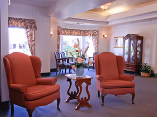 A cozy sitting area in a senior living facility with two red upholstered armchairs facing each other and a small wooden table with a floral arrangement in between. In the background, there are large windows with floral curtains, a wooden china cabinet, and additional seating near the windows.