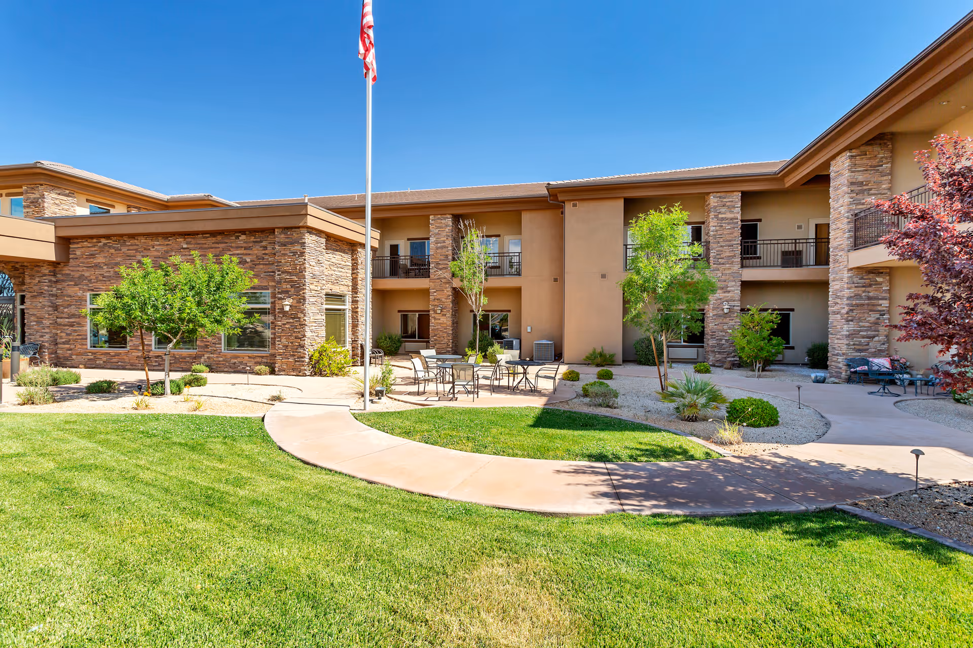 Outdoor courtyard area of a senior living facility with green grass, paved walkways, patio tables and chairs, small trees, and a two-story building with stone and stucco exterior under a clear blue sky.