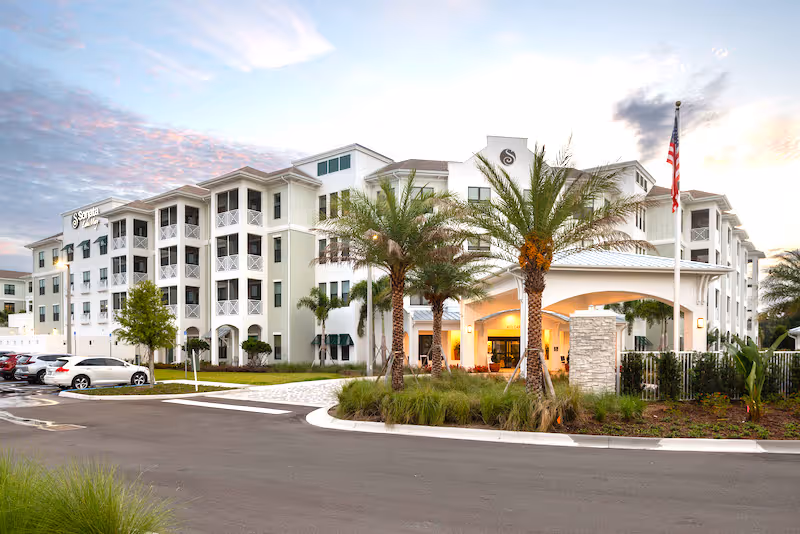 Exterior front view of the Sonata Lake Mary senior living building with a covered entrance, palm trees, and parked cars.