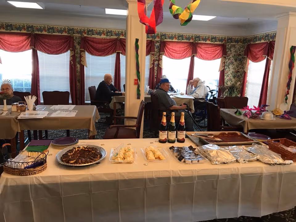 Decorated dining room with a long buffet table of food and drinks and several elderly residents seated at tables near windows.