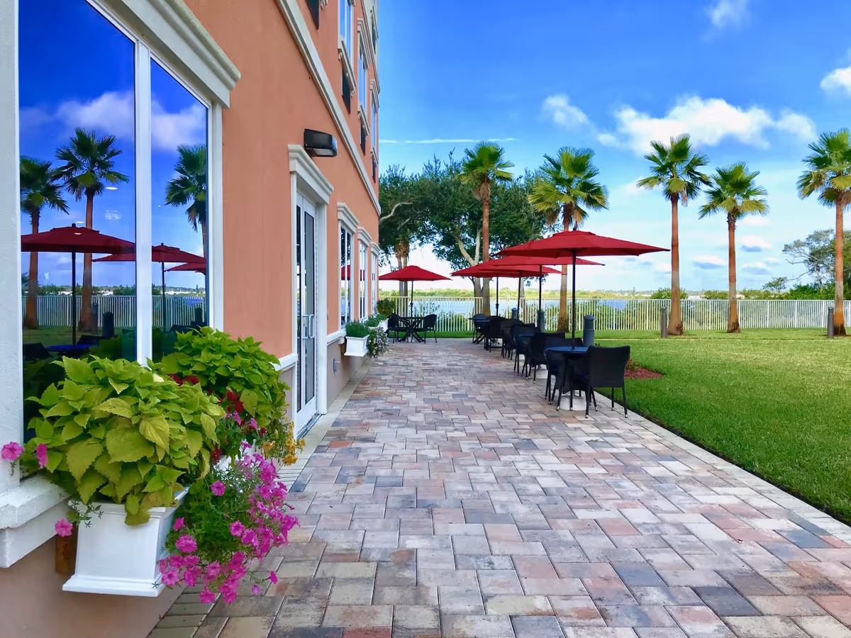 Outdoor patio area at Heritage Waterside Luxury Senior Living with a paved walkway, black tables and chairs under red umbrellas, palm trees, green lawn, and a view of a lake in the background.