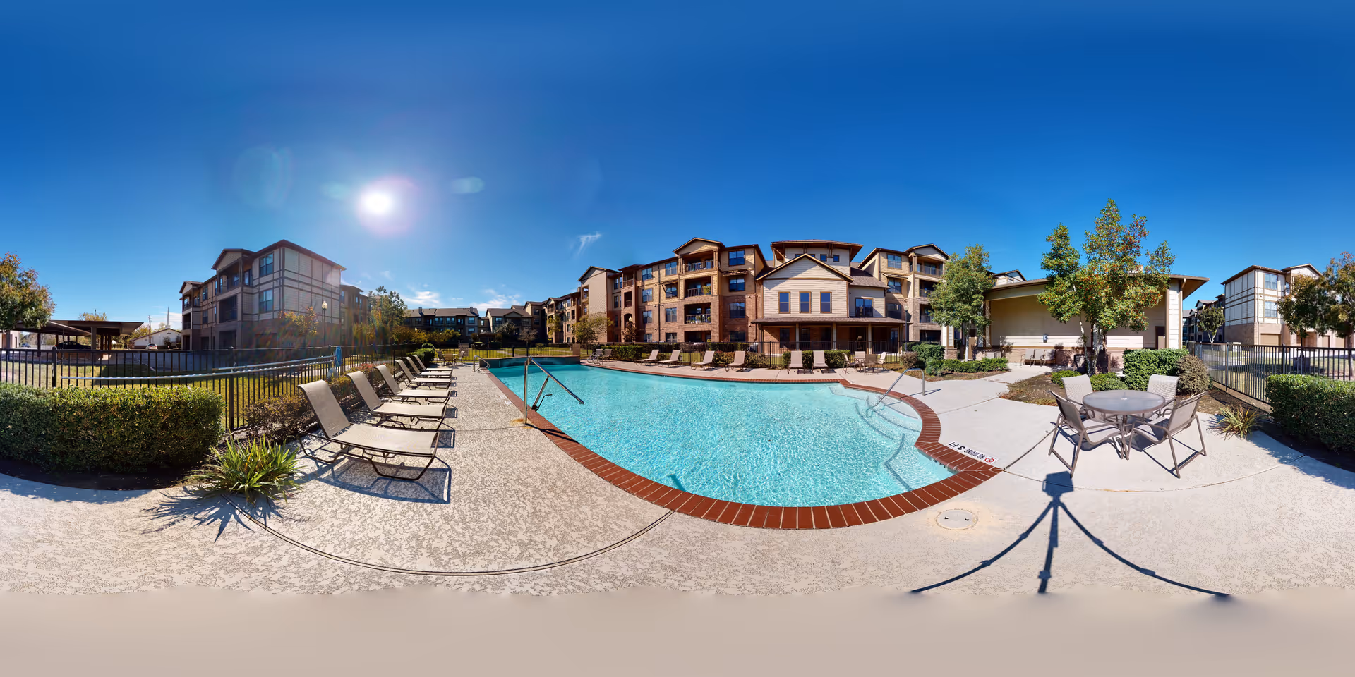 Outdoor swimming pool area at a senior living facility with lounge chairs lined up along the poolside, a round table with chairs, surrounded by multi-story residential buildings and trees under a clear blue sky.