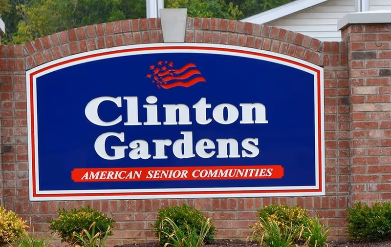 A large blue and white sign with red accents mounted on a brick wall. The sign reads 'Clinton Gardens' with 'American Senior Communities' written below in red. There are small green bushes and plants in front of the brick wall.