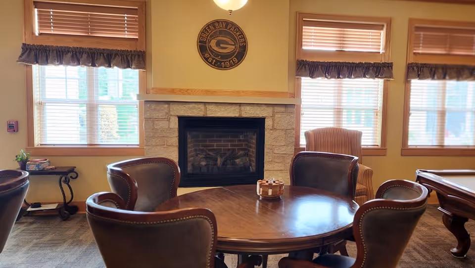 A cozy common area with a round wooden table surrounded by five leather chairs. Behind the table is a stone fireplace with a Green Bay Packers sign above it. The room has three windows with wooden blinds and valances, and there is a striped armchair near the windows. A small side table with board games is visible to the left.