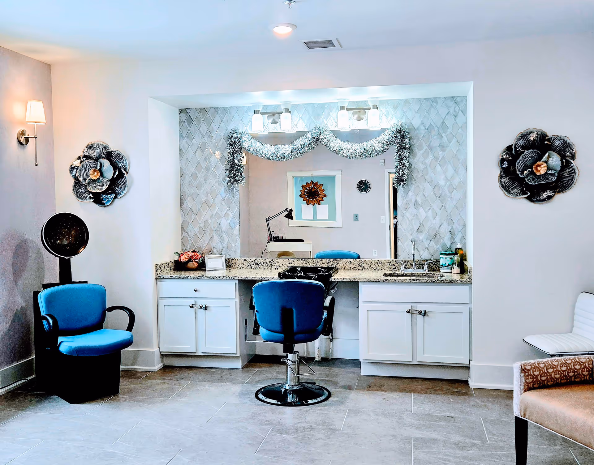 Interior view of a salon area in a senior living facility with a blue salon chair in front of a large mirror decorated with silver garland. The room has white cabinets, a granite countertop, a hair dryer chair on the left, and decorative flower wall art on both sides of the mirror.