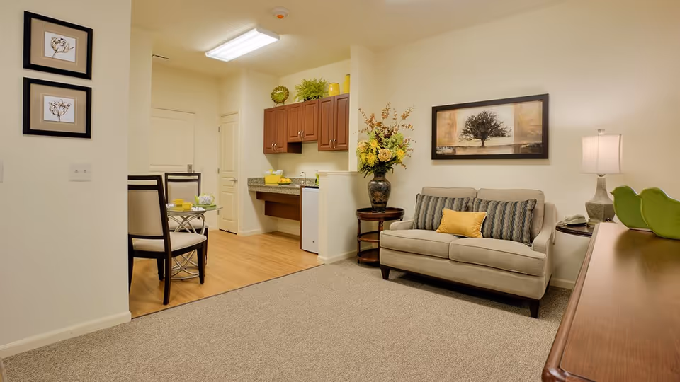 A cozy living area in an assisted living facility featuring a beige sofa with striped and yellow cushions, a side table with a lamp, a large floral arrangement on a round table, and a framed picture of a tree on the wall. Adjacent to the living area is a small dining space with a glass-top table and two chairs, and a kitchenette with wooden cabinets, a countertop, and a mini refrigerator. The room has light-colored walls and a combination of carpet and wood flooring.