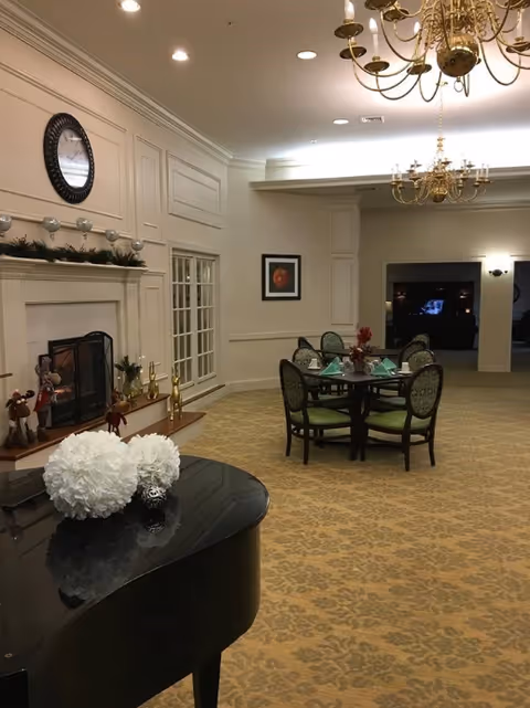 Common area in a senior living facility with a grand piano, fireplace, and a round dining table under chandeliers.
