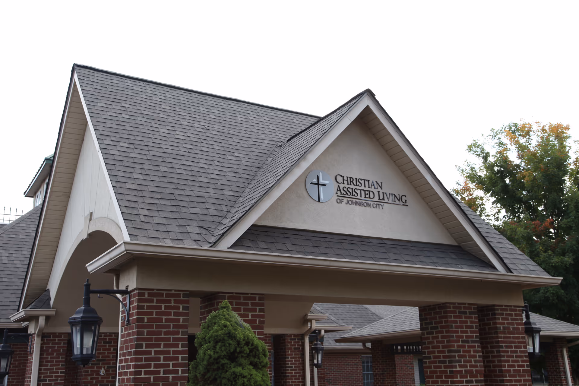 Entrance of a building with a peaked roof and brick pillars, displaying a sign that reads 'Christian Assisted Living of Johnson City' with a cross logo. There are outdoor lantern-style lights attached to the pillars and some trees in the background.
