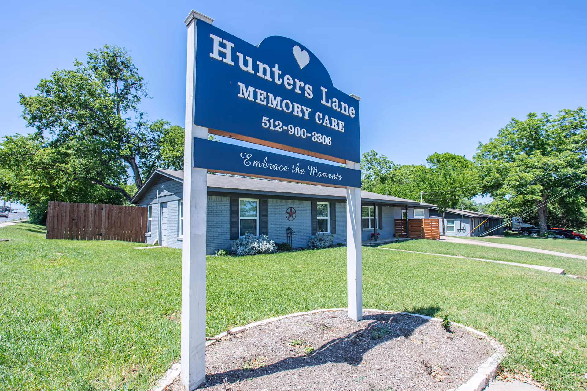 Blue sign reading 'Hunters Lane Memory Care' in front of a single-story building, lawn, and trees under a clear sky.