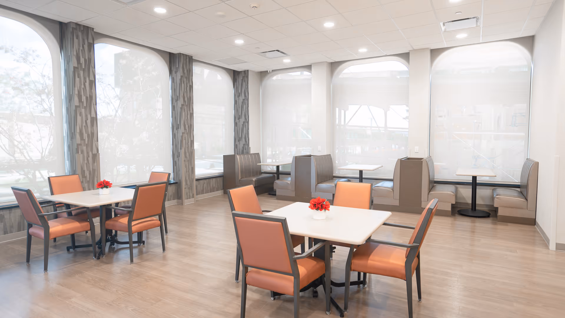 A bright dining room with large arched windows covered by translucent blinds. The room features several tables with orange cushioned chairs and booth seating along the windows. Each table has a small red flower centerpiece. The floor is light wood, and the walls are light-colored with patterned accents between the windows.