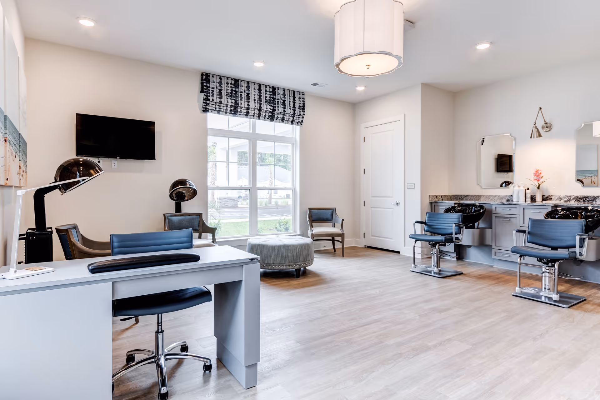 A bright and modern salon area with two blue salon chairs in front of black wash basins, a desk with a blue chair, two hair drying stations with chairs, a large window with patterned valance, and light wood flooring.