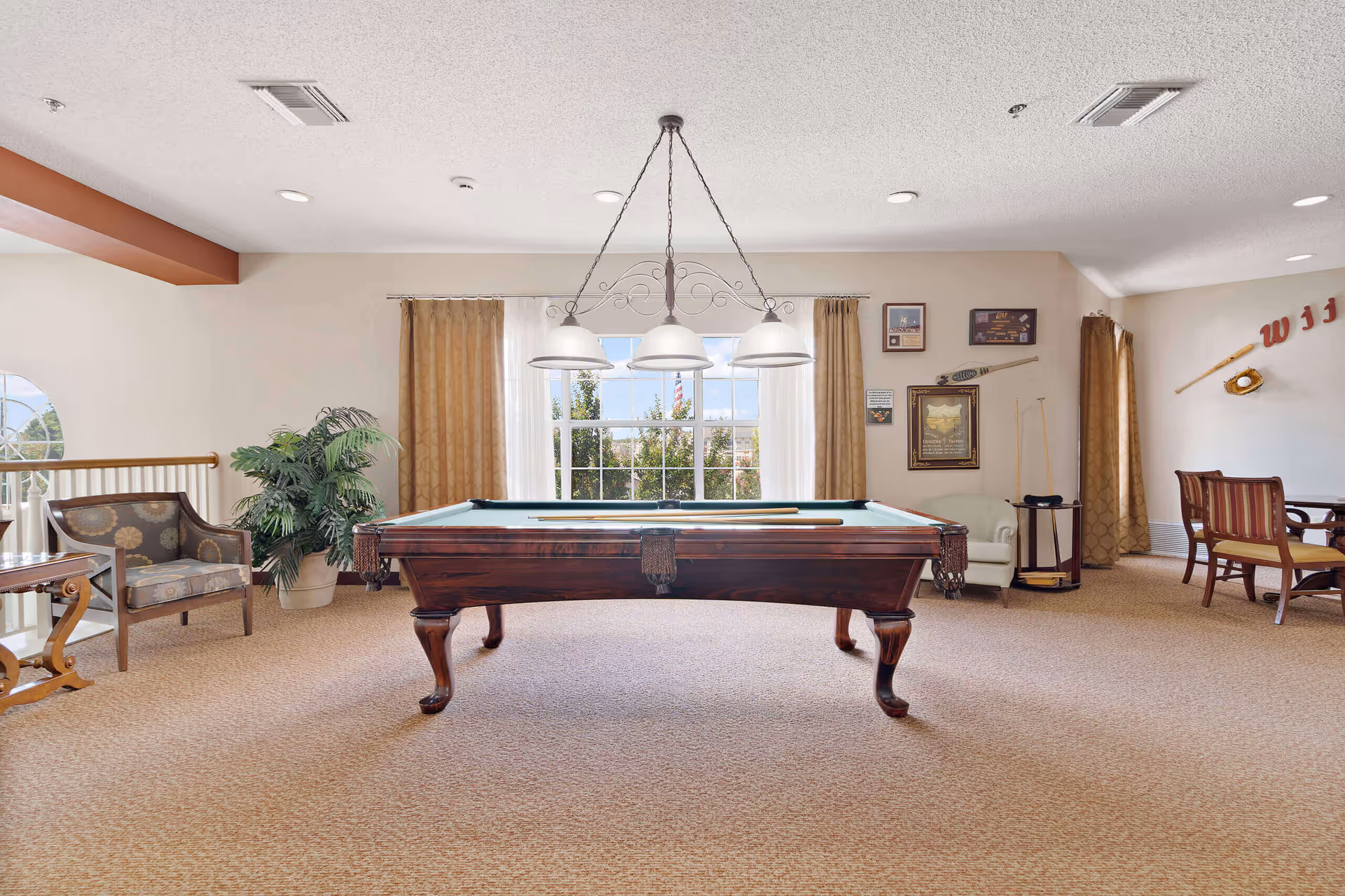 A well-lit recreational room with a pool table in the center, a hanging light fixture above it, a large window with beige curtains in the background, and seating areas with chairs and a small table on the sides. The room has beige carpet and light-colored walls with some framed pictures and decorations.