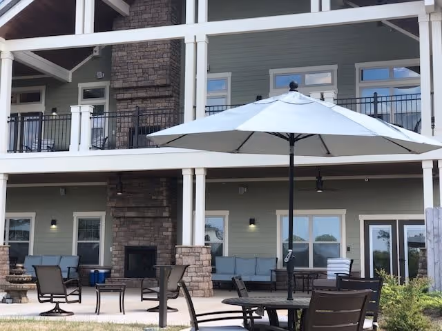 Outdoor patio area at a senior living facility with multiple seating arrangements including chairs and tables. There is a large white umbrella providing shade over one table. The building behind has a stone fireplace and multiple windows and doors, with a balcony above.