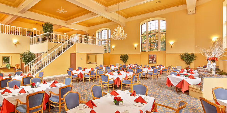 Spacious dining room with multiple tables covered in white and red tablecloths, each set with cups, saucers, and red folded napkins. The room features high ceilings, large arched windows, a chandelier, and a staircase leading to an upper level. There are plants and framed artwork on the walls, and upholstered chairs with wooden frames around the tables.