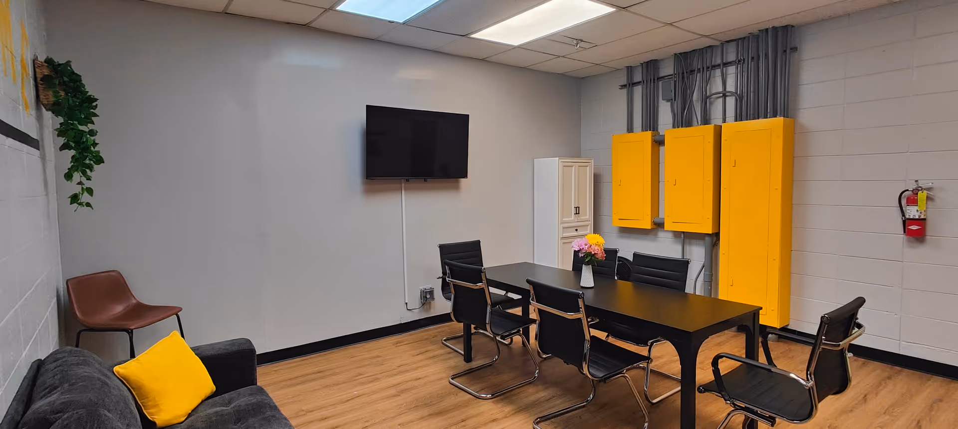 Small communal meeting room with a black table and chairs, a wall-mounted TV, a gray sofa with a yellow pillow, and bright yellow wall cabinets.