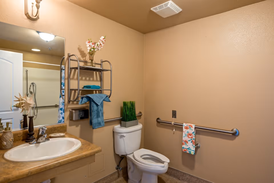 A clean bathroom with beige walls featuring a white toilet with a floral towel hanging on a metal grab bar beside it. Above the toilet is a metal shelf holding a vase with flowers and folded blue towels. To the left is a sink with a brown countertop, a soap dispenser, and decorative items. A mirror and a partially open door leading to a shower area are also visible.