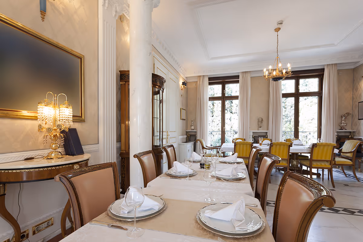 Elegant dining room with a long table set for a meal, featuring white tablecloths, folded napkins, wine glasses, and silver plates. The room has large windows with white curtains, classic wooden chairs with leather upholstery, ornate wall decorations, a chandelier, and a decorative lamp on a side table.