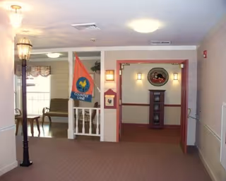 Interior hallway of a senior living facility with beige walls and carpet. On the left side, there is a decorative street lamp and a small white railing with a colorful flag that reads 'Country Lane.' Beyond the railing, there is a seating area with chairs and a table near a window with curtains. On the right side, there is an open doorway with red trim leading to a small alcove featuring a tall wooden grandfather clock and a round wall decoration above it. Ceiling lights illuminate the space.