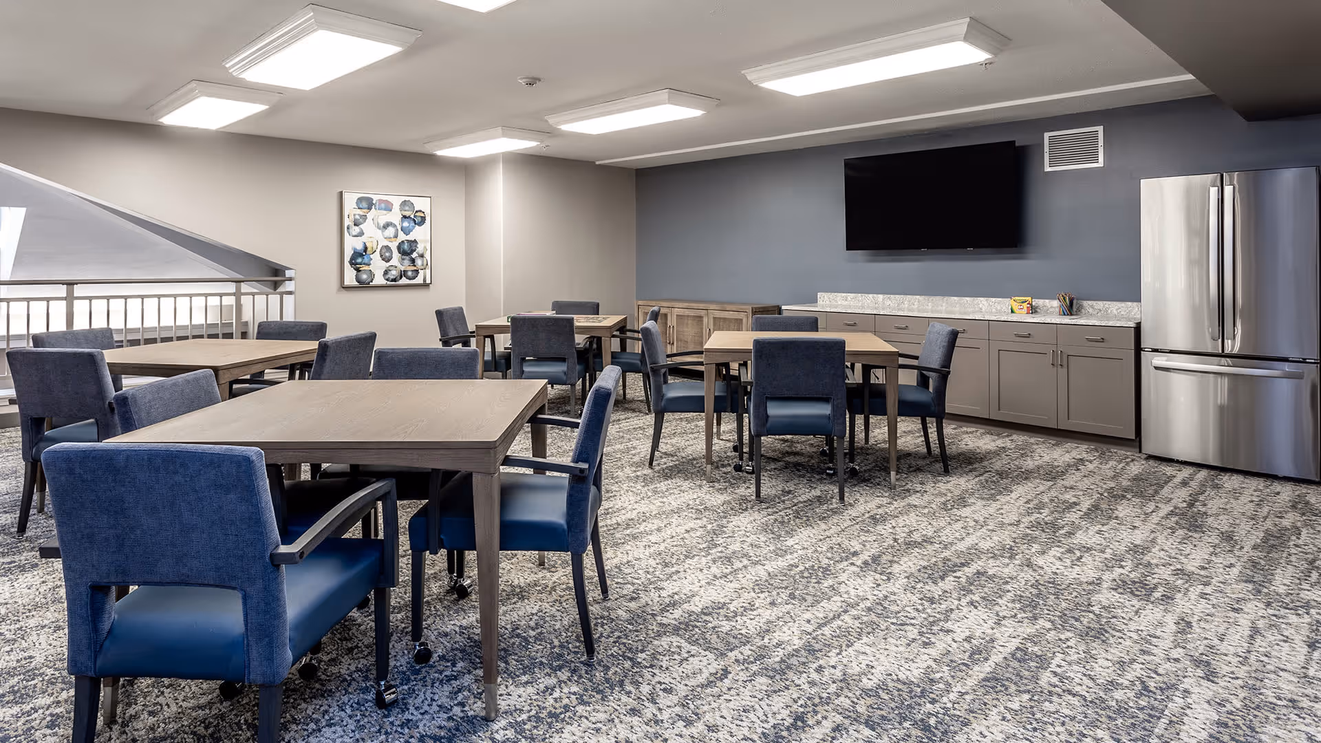 A modern common room with several wooden tables and blue upholstered chairs arranged neatly on a patterned carpet. The room features a large flat-screen TV mounted on a gray wall, a stainless steel refrigerator, and gray cabinetry with a marble countertop. There is a piece of abstract art on one wall and bright overhead lighting.