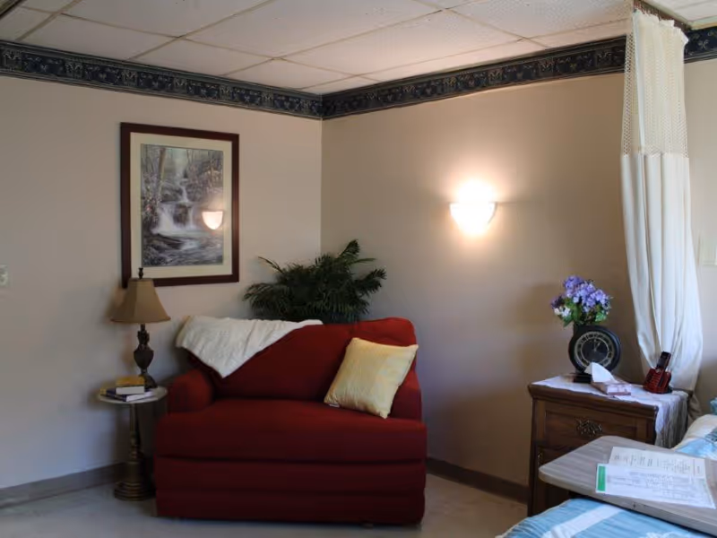 A cozy corner of a room featuring a red armchair with a yellow pillow and a white throw blanket. Next to the chair is a small round table with a lamp and some books. On the wall above the chair is a framed picture of a waterfall. To the right, there is a wooden nightstand with a clock, a vase of purple flowers, and some papers. A white curtain hangs partially visible on the right side of the image.