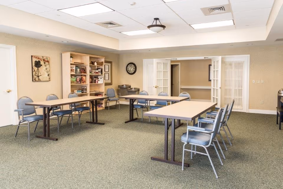 A well-lit meeting or activity room with two rectangular tables arranged in a U-shape surrounded by blue cushioned chairs. The room has beige walls, a green carpet, a wall clock, a wooden bookshelf filled with books and decorative items, and double glass doors leading to another room.