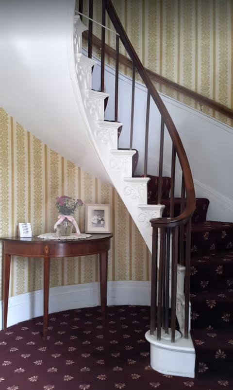 Curved wooden staircase with white decorative trim and dark wooden handrail in a room with patterned wallpaper and carpet. A small wooden table with a lace doily, a vase of flowers, and framed photos is placed against the wall under the staircase.
