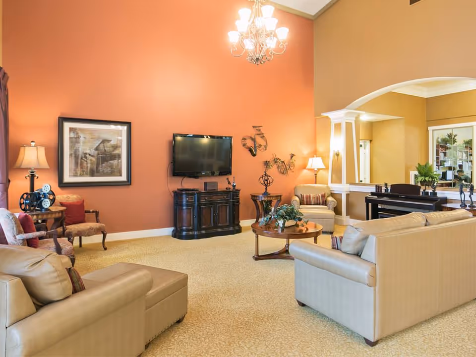 A spacious living room with beige leather sofas, a round wooden coffee table with a plant centerpiece, and a flat-screen TV mounted on a coral-colored wall. The room features a chandelier, framed artwork, decorative metal wall art, a piano, and several lamps providing warm lighting. The carpet is light-colored with a subtle pattern, and there is an archway leading to another room with bookshelves.