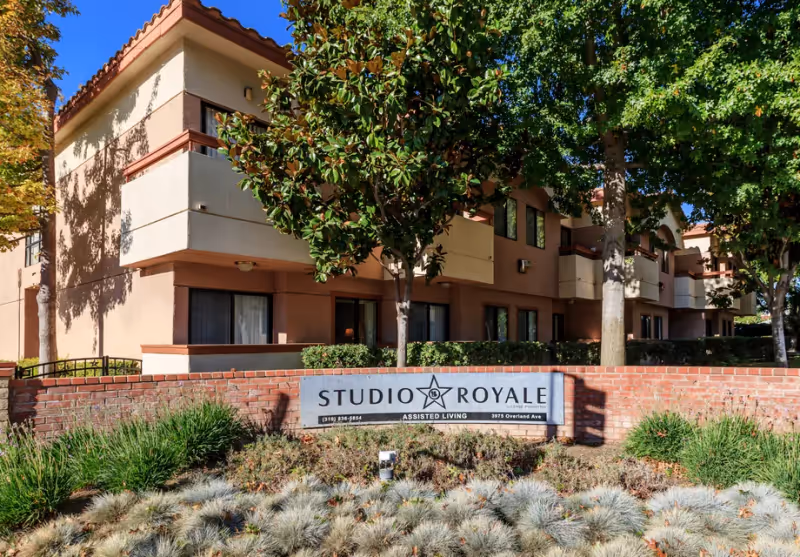 Front exterior of the Studio Royale assisted living building with a brick sign and landscaped shrubs.