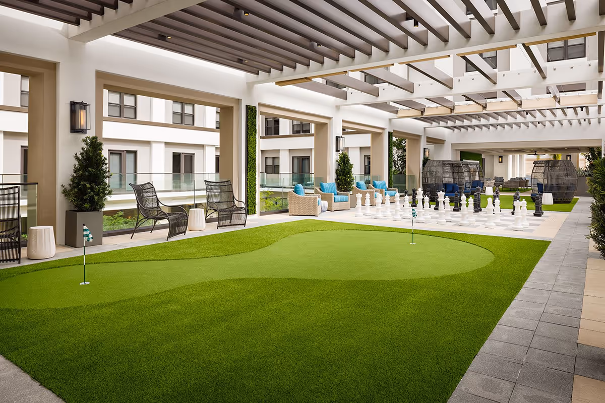 Covered outdoor recreational area with artificial putting green, large chess set, wicker chairs with blue cushions, and modern black chairs. The space is surrounded by a building with large windows and has a pergola-style ceiling.