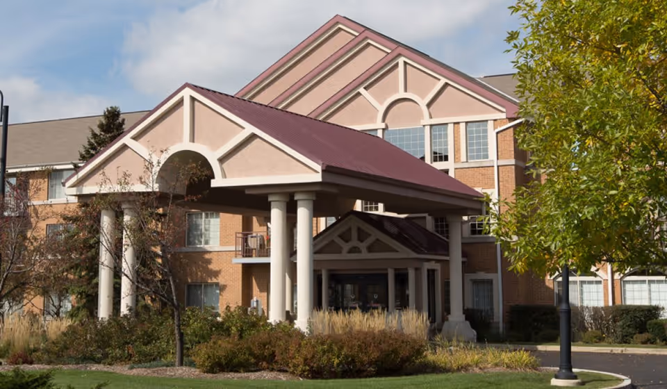 Exterior view of a senior living facility building with a covered entrance supported by white columns, surrounded by trees and landscaping under a partly cloudy sky.