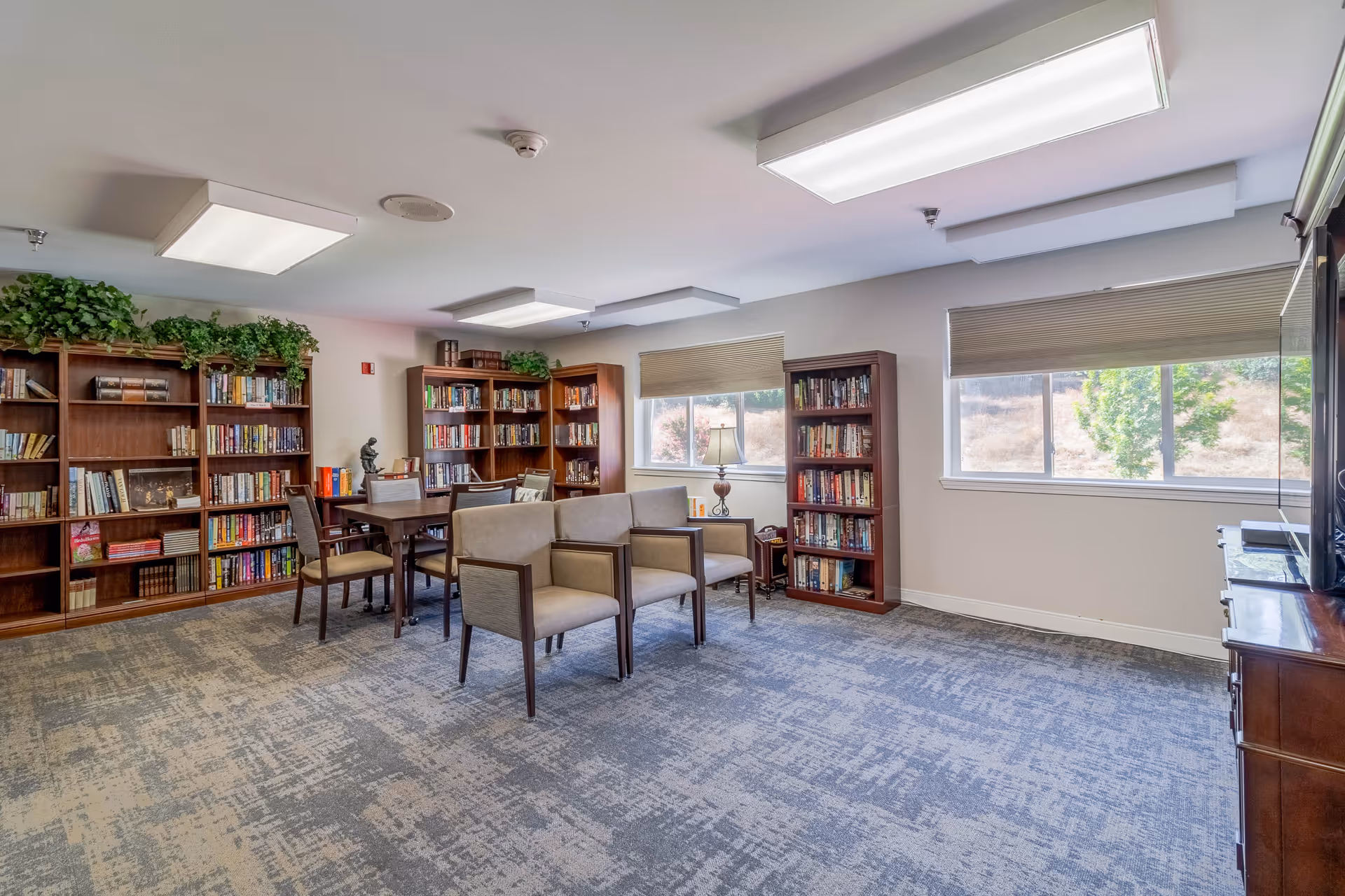 A bright and spacious library or reading room in a retirement community with multiple wooden bookshelves filled with books, a table with chairs, and two armchairs facing a television. The room has large windows with beige blinds, a patterned carpet, and ceiling lights.