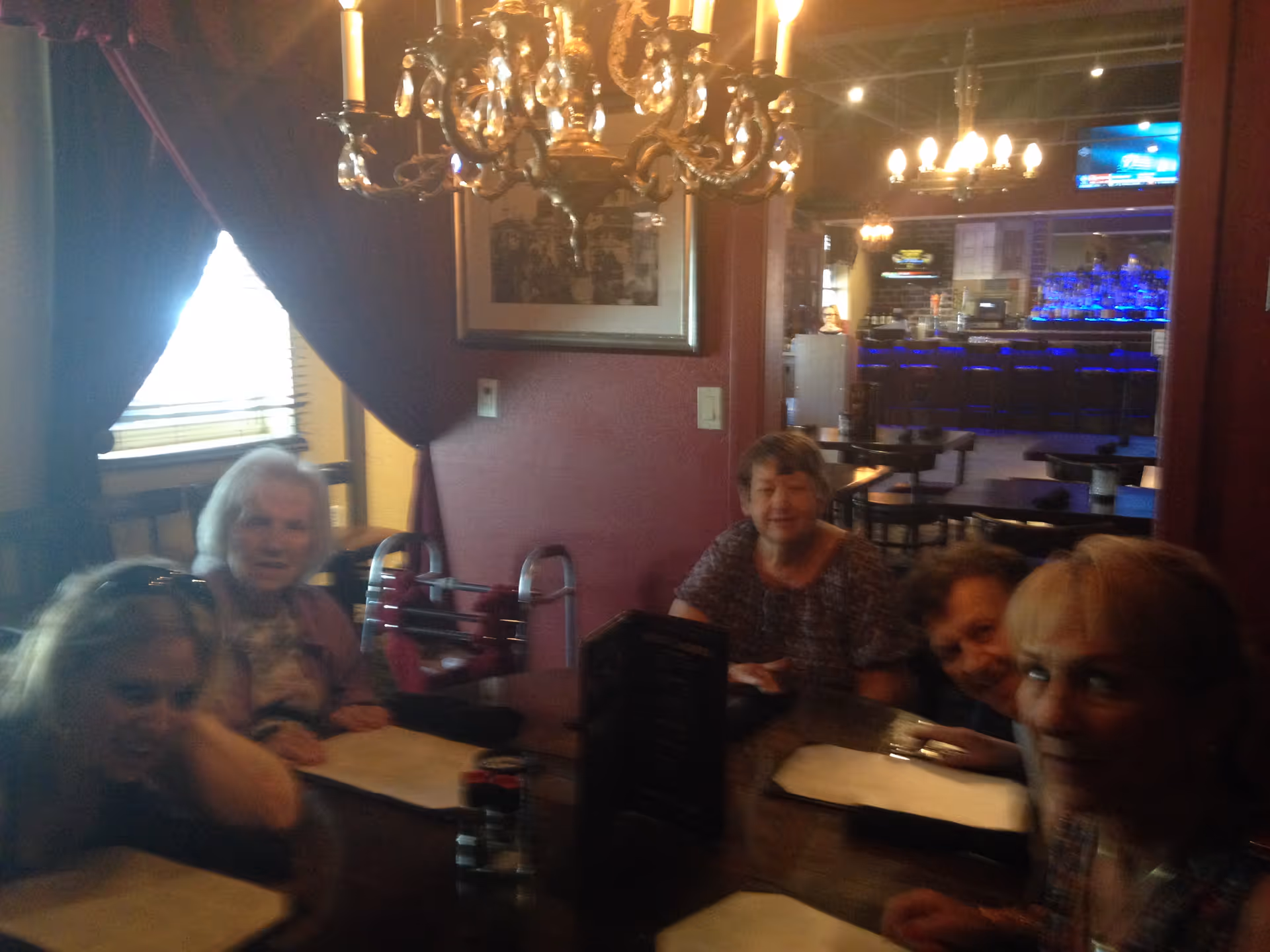 A group of older adults seated around a table in a dimly lit dining area with a chandelier and a bar visible in the background.