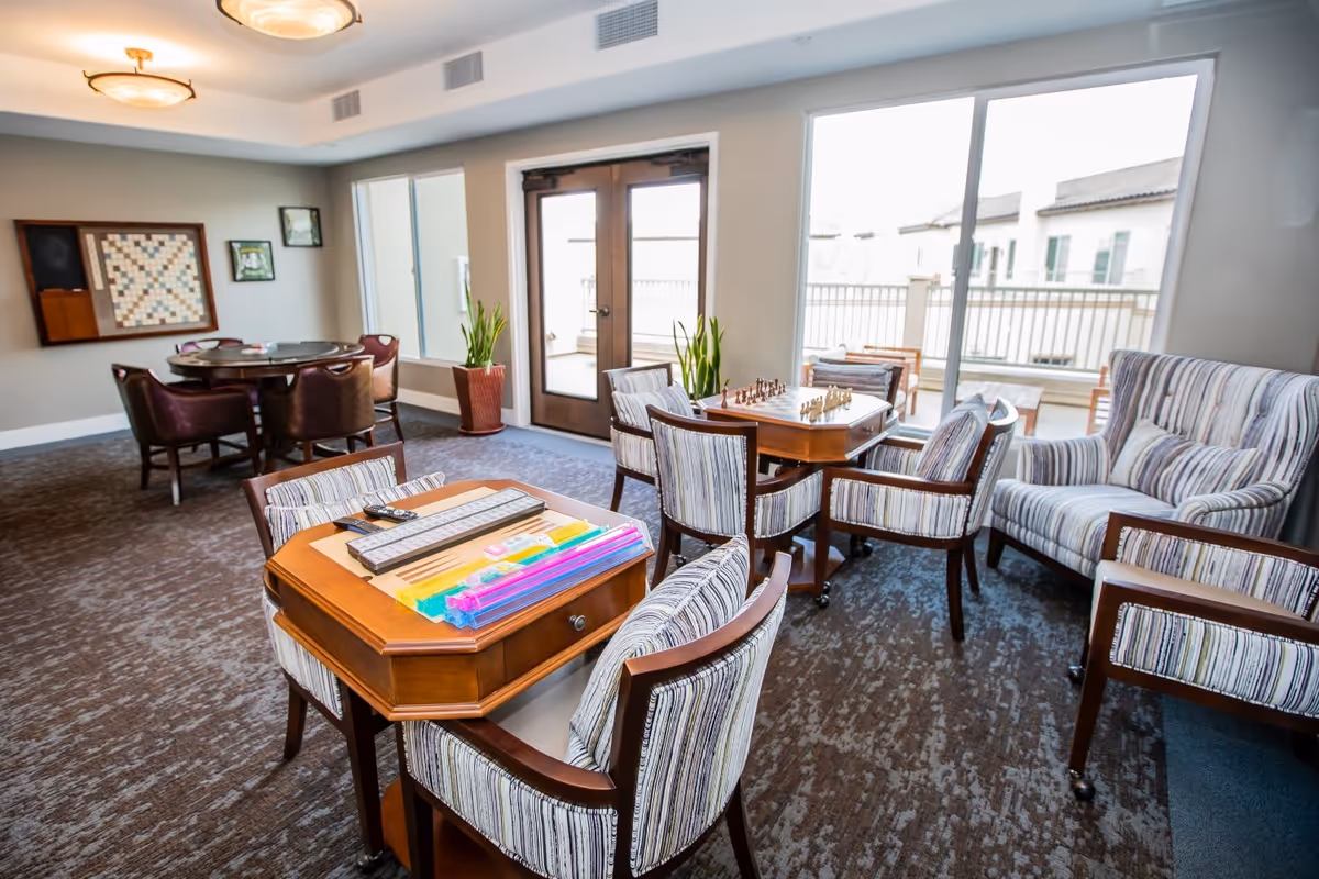 Bright communal lounge with tables, striped armchairs, a chessboard and large windows opening to a balcony.