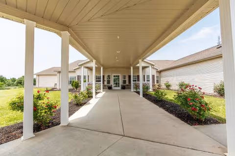 Covered drive-up entrance with white columns and landscaped flower beds leading to the front doors of a single-story senior living facility.