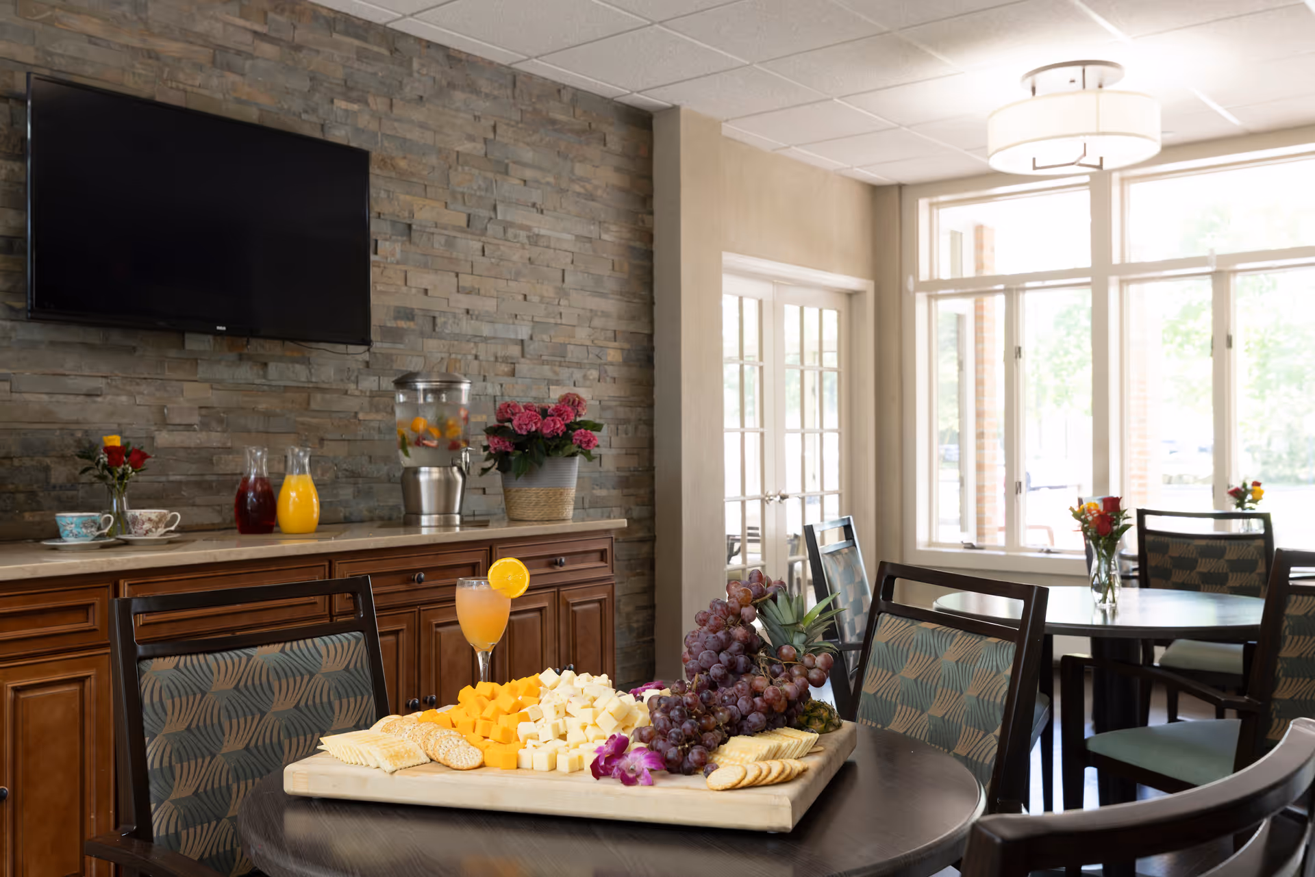 A dining area with round tables and chairs featuring patterned upholstery. On one table, there is a wooden board with an assortment of cheese cubes, crackers, grapes, and a cocktail garnished with an orange slice. In the background, there is a wooden cabinet with a stone wall behind it, holding a beverage dispenser with fruit-infused water, two pitchers of juice, and a potted plant with pink flowers. A flat-screen TV is mounted on the stone wall above the cabinet. Large windows and glass doors allow natural light to fill the room.
