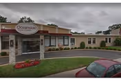 Exterior view of Oceanside Nursing Center showing the entrance with a covered drop-off area, a sign with the facility name, landscaped greenery, and a parked red car in the driveway under a cloudy sky.