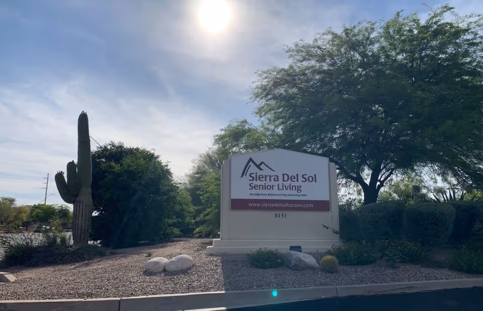 Outdoor view of the Sierra Del Sol Senior Living facility sign surrounded by desert landscaping including a large cactus, bushes, and trees under a partly cloudy sky with the sun shining.