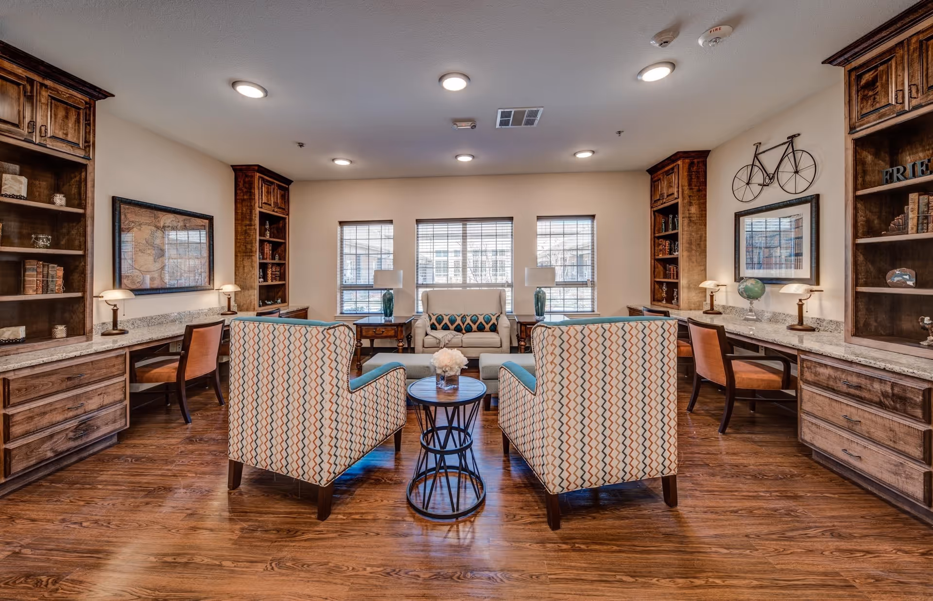 A cozy and well-lit common area with wooden flooring, two patterned armchairs facing a small round table with a flower arrangement, a loveseat with decorative pillows, built-in wooden bookshelves and desks with chairs on either side, and three windows letting in natural light.