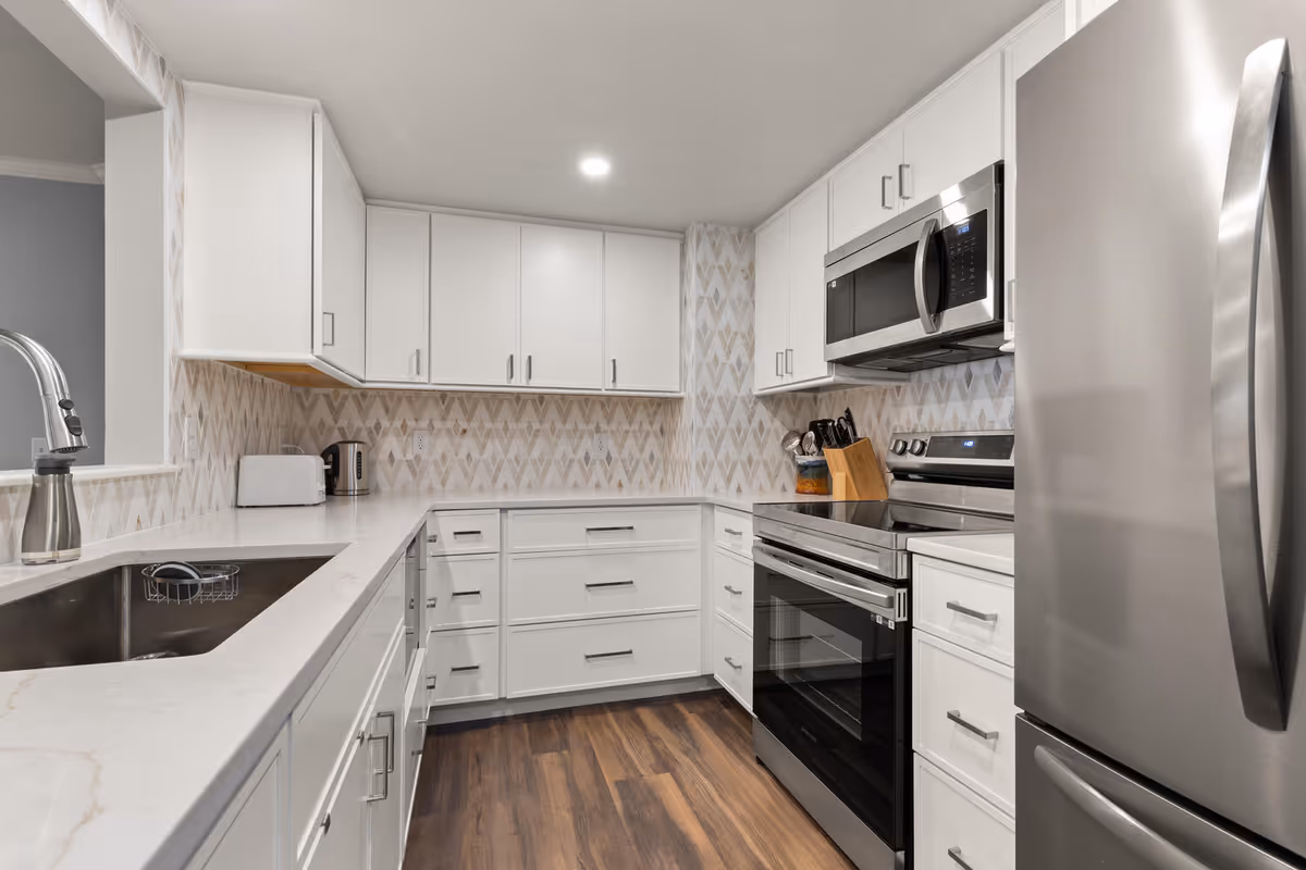 Modern kitchen with white cabinets, a stainless steel refrigerator, oven, microwave, and a sink with a curved faucet. The backsplash features a geometric pattern, and the floor is wood.