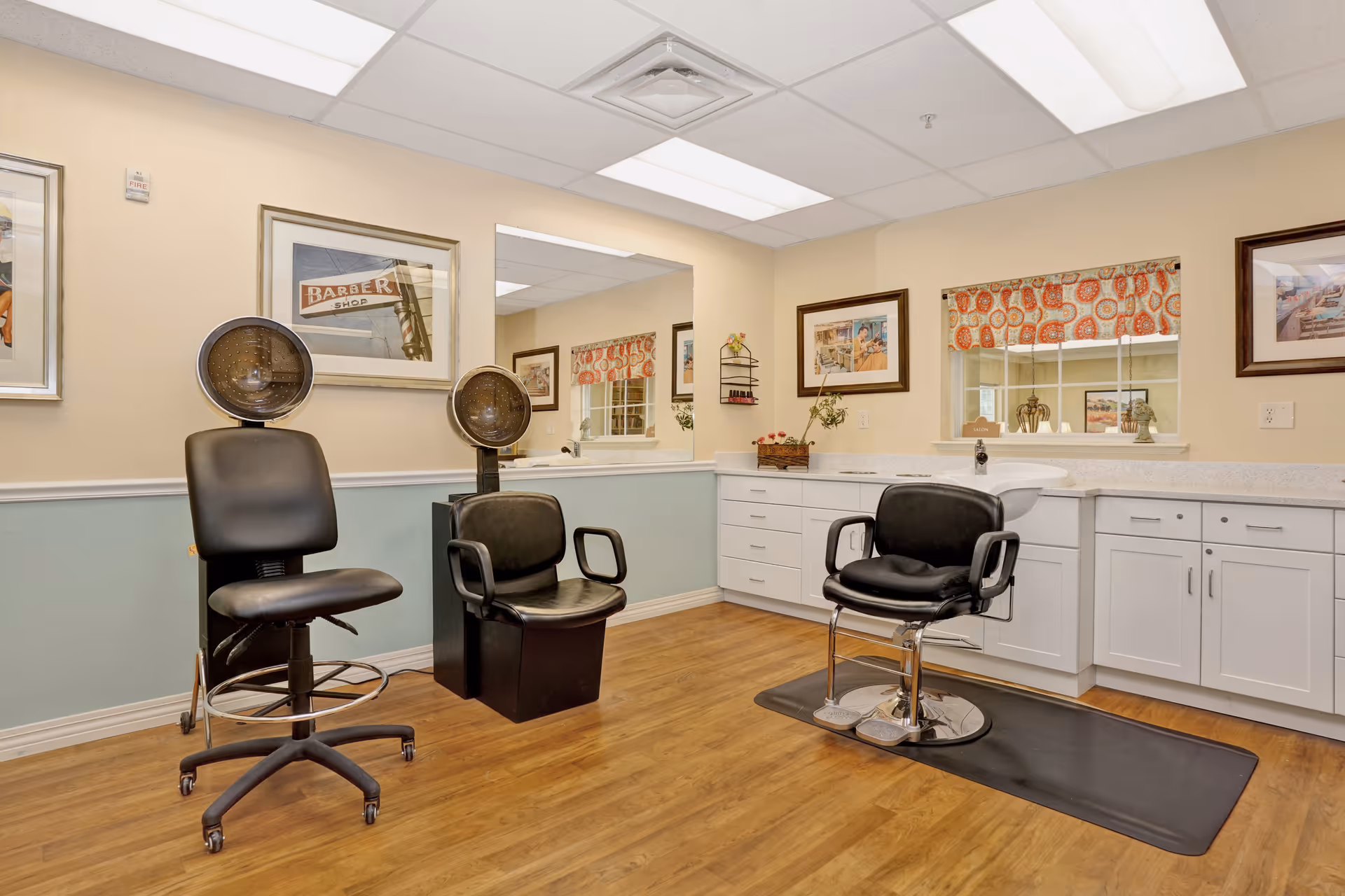 Interior of a salon area in a senior living facility with three black salon chairs, two hair dryers, white cabinets with countertop, framed pictures on the walls, a window with patterned curtains, and wood flooring.