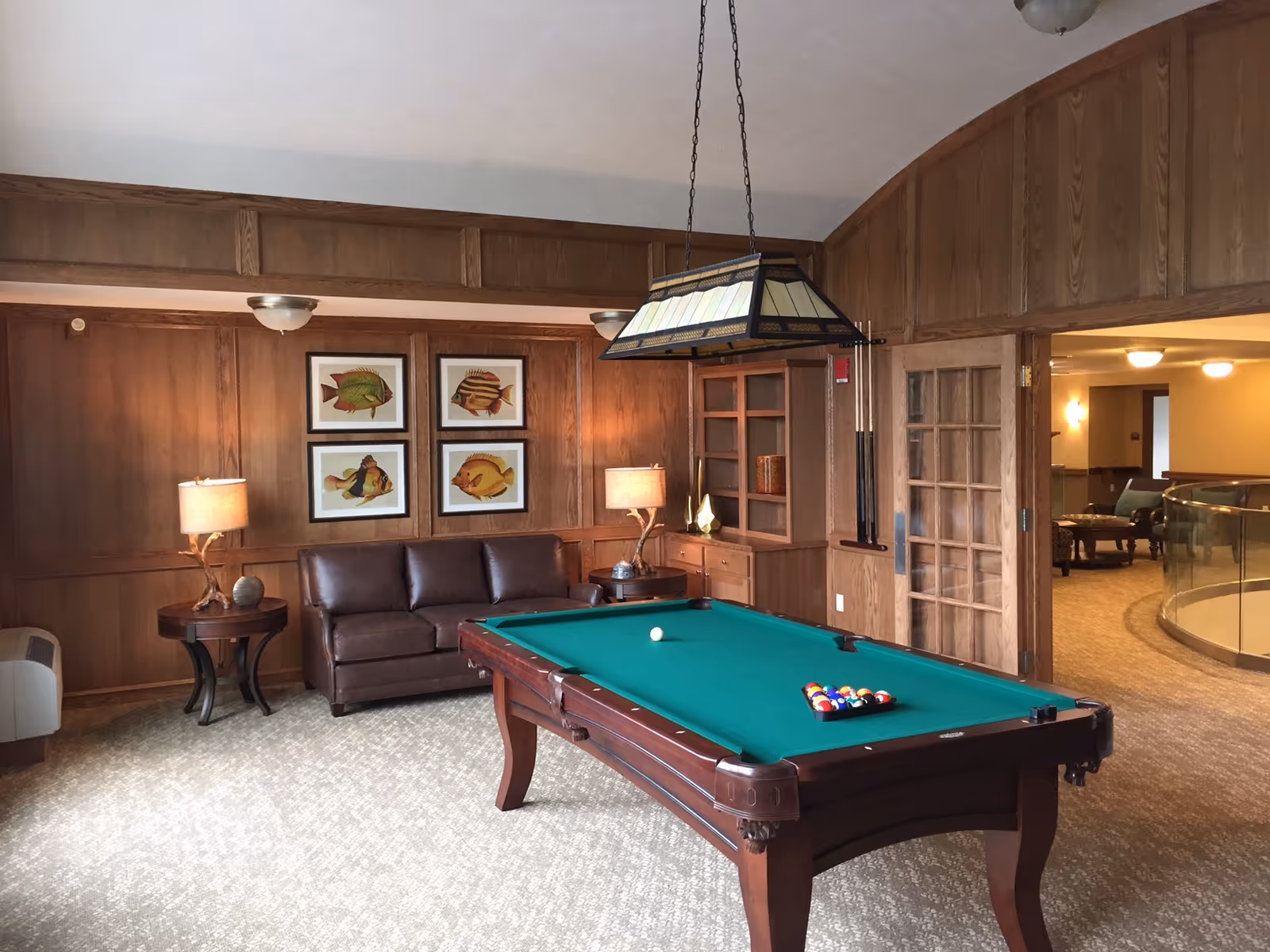 A cozy game room with a green felt pool table in the center, a brown leather couch against a wood-paneled wall, two side tables with lamps, and four framed fish illustrations hanging above the couch. There is a hanging light fixture above the pool table and a doorway leading to another room with seating and a curved glass railing.
