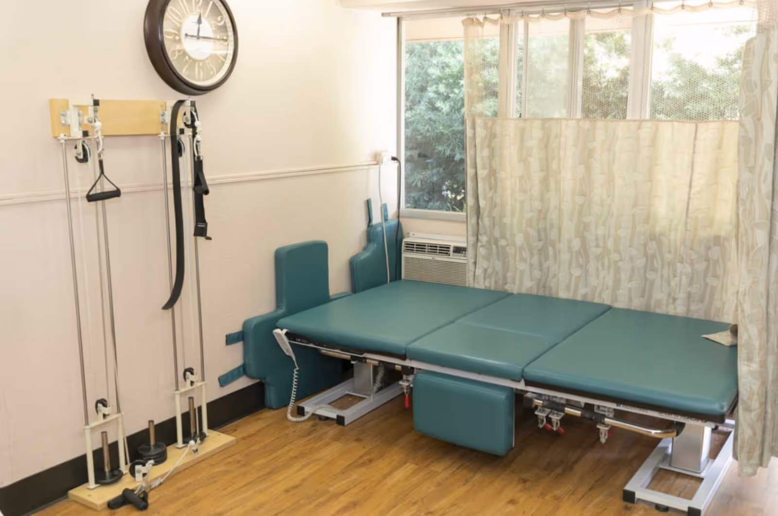 A physical therapy room with a padded treatment table, exercise pulleys mounted on the wall, a large clock, and a window with curtains partially drawn. The room has wooden flooring and light-colored walls.