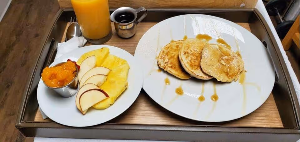 Tray holding a plate of pancakes drizzled with syrup, a side plate with pineapple and apple slices plus a fruit cup, a small pitcher of syrup and a glass of orange juice.